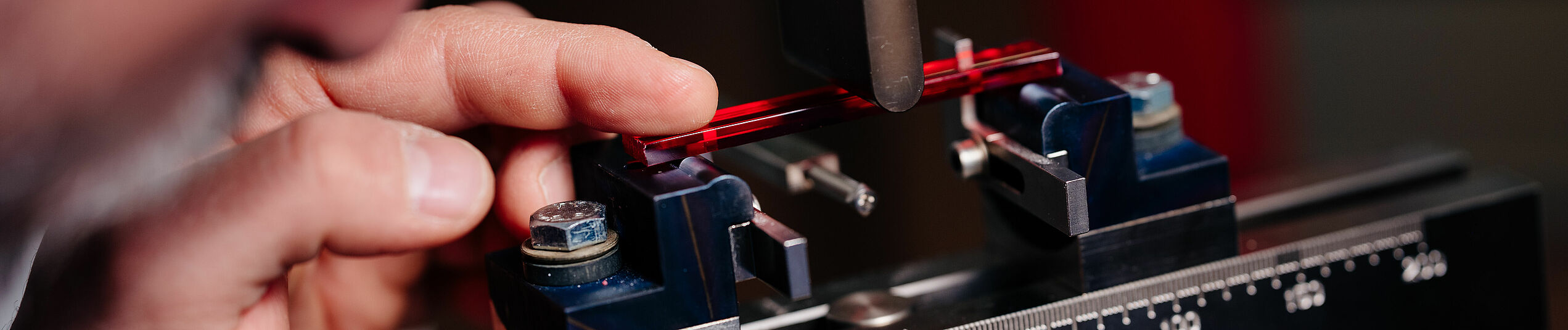 Ein Wissenschaftler bereitet die Messung der Durchbiegefestigkeit eines rot farbigen transparenten Kunststoffstreifens in einer Apparatur vor.