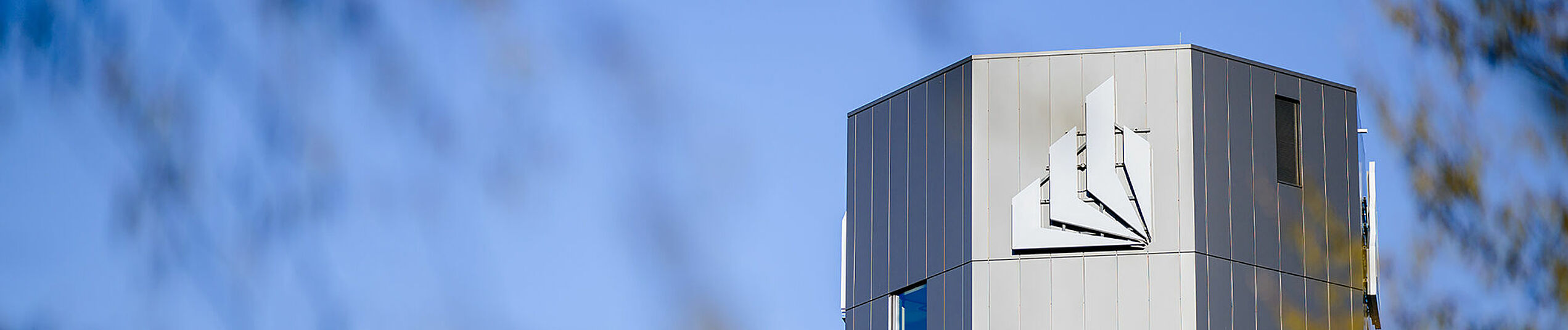 Der obere Teil des dunkelgrauen oktaedrischen Turm mit der Bildmarke des Logos der Uni Paderborn vor blauem Himmel.
