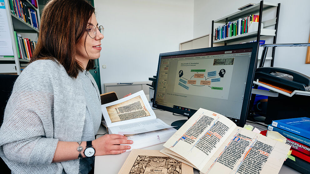 Eine Wissenschaftlerin mit einem aufgeschlagenen Buch, dass links die Abbildung eines mittelalterlichen Schriftstcks enth?lt, daneben zwei weitere Bcher. Dahinter ein Widescreen-Monitor mit der Anzeige einer Vortragsfolie "Maschinelle CC-Annotation"