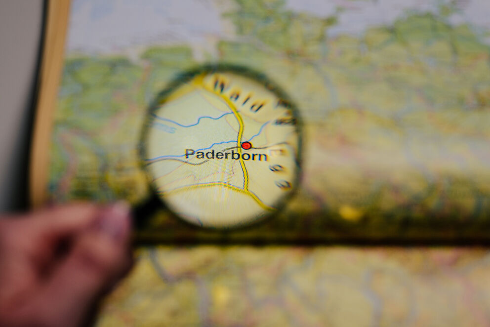 Eine Hand h?lt eine Lupe vor einen Atlas, die Stadt Paderborn befindet sich im Zentrum der Lupe und wird hervorgehoben.